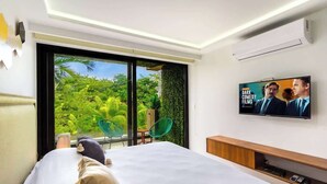 Ein modernes Hotelzimmer mit einem großen Bett, einem an der Wand montierten Fernseher und einer Schiebetür zu einem Balkon mit Blick auf Grünflächen.