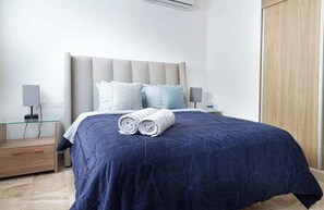 Una cama bien hecha con colcha azul oscuro, dos almohadas y toallas enrolladas sobre una mesita de noche de madera.