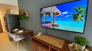 A modern living room with a flat-screen TV mounted on the wall, a wooden coffee table, a dining area with a small table and chairs, and a potted plant on the coffee table. - Studio w Rooftop Pool Near Beach & 5th Ave (Playa del Carmen)