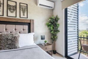 A bedroom with a tufted headboard, a bed with a fluffy pillow, a bedside table with a lamp and a small plant, and a large window with blinds. - Studio w Rooftop Pool Near Beach & 5th Ave (Playa del Carmen)