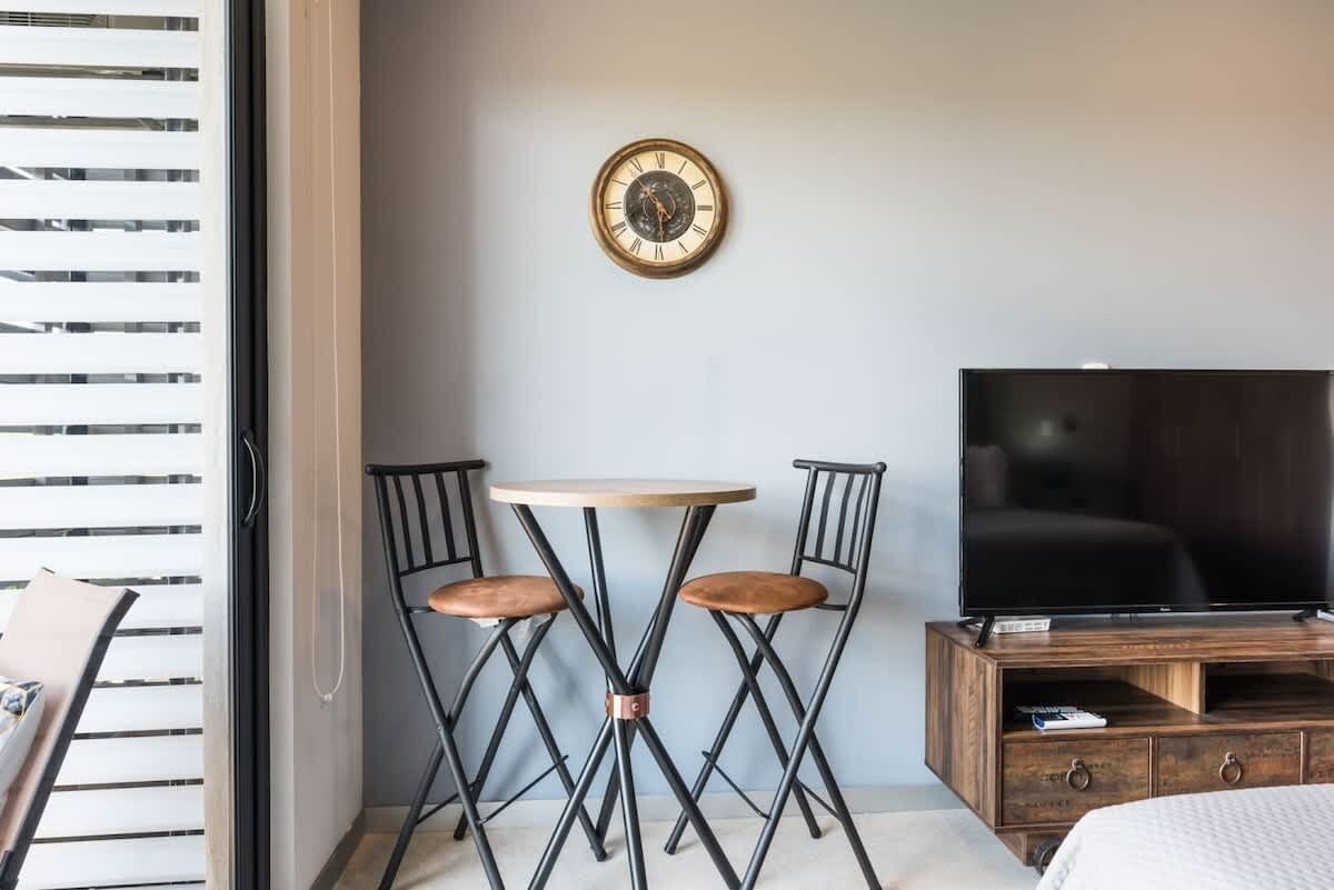 Una habitación pequeña con una mesa redonda y dos sillas, un televisor sobre un mueble de madera y un reloj de pared.