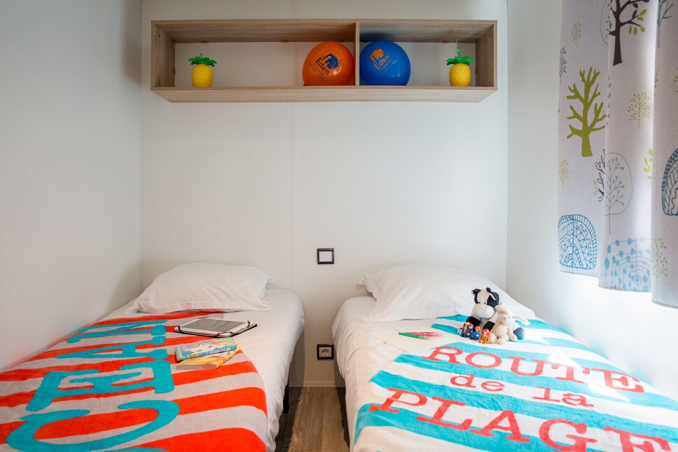 A bedroom with two beds, each with a colorful bedspread, a shelf above with decorative items, and a window with curtains.