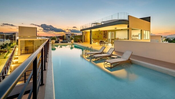 Una piscina en la azotea con sillones, barandilla de cristal y un edificio moderno con ventanales grandes.