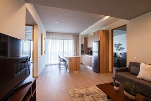 Una sala moderna con un televisor de pantalla plana, un sofá y una mesa de centro. Cuenta con una zona de cocina con refrigerador y una zona de comedor con taburete de bar.