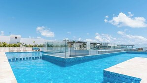 A modern swimming pool with glass walls, a clear blue sky, and a white building in the background. - Rooftop Pool and Private Hot Tub with Ocean Views (Playa del Carmen)