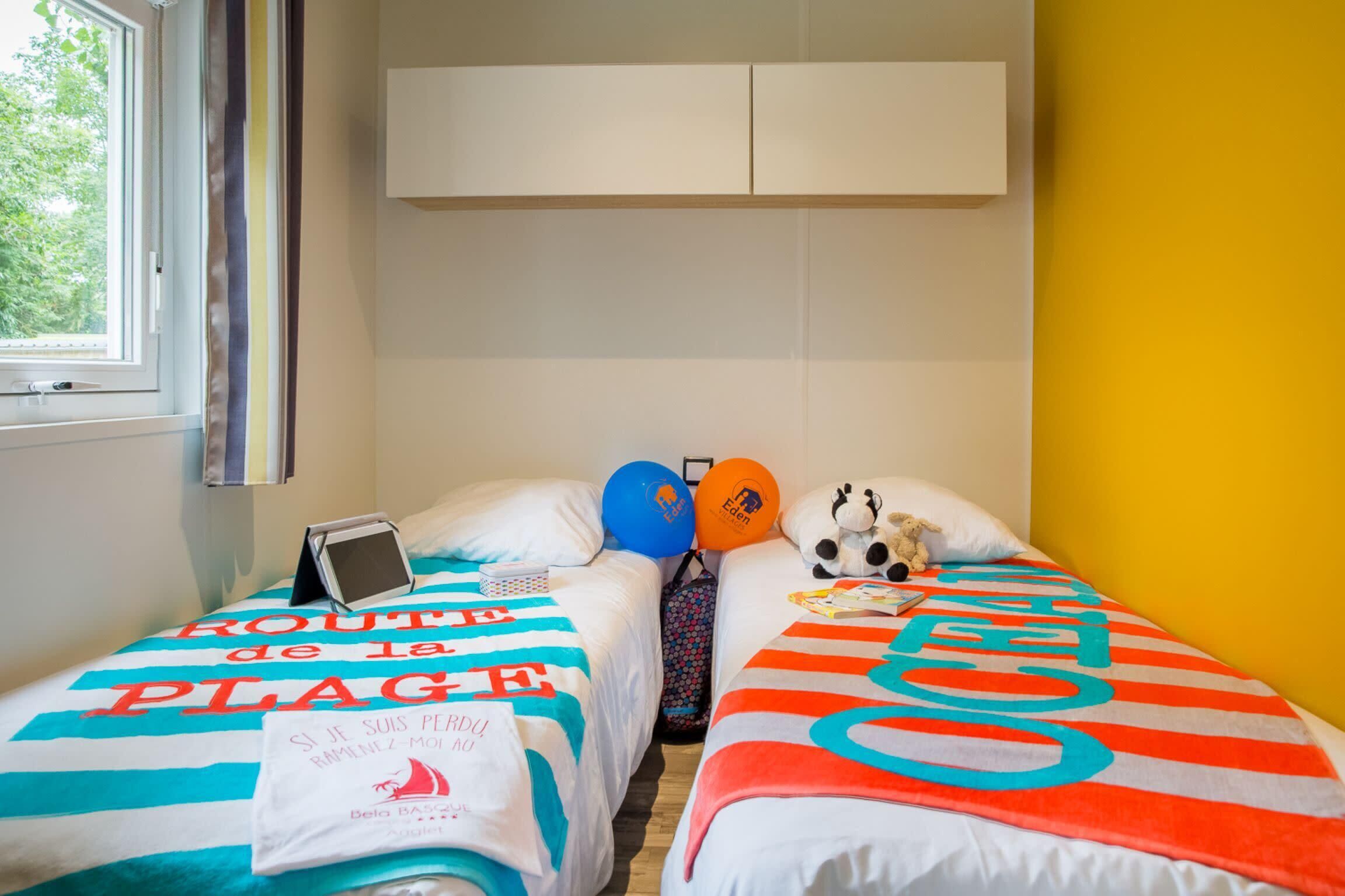 Une chambre avec deux lits, chacun recouvert d’une courtepointe colorée. Il y a une fenêtre sur la gauche, une armoire blanche au-dessus des lits et un mur jaune sur la droite.