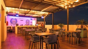 An outdoor dining area with wooden tables and chairs, a bar with a neon sign, and a view of the night sky. - Modern Studio w Rooftop Pool Near Beach & Dining (Playa del Carmen)