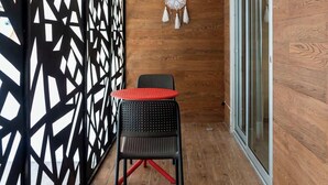 A small, enclosed balcony with a wooden floor, a black and white patterned partition, a small table with a red top, two black chairs, and a decorative item on the wall. - Modern Studio w Rooftop Pool Near Beach & Dining (Playa del Carmen)