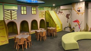 Zona de juegos infantiles con paredes de escalar, tobogán y área de descanso con mesas y sillas.