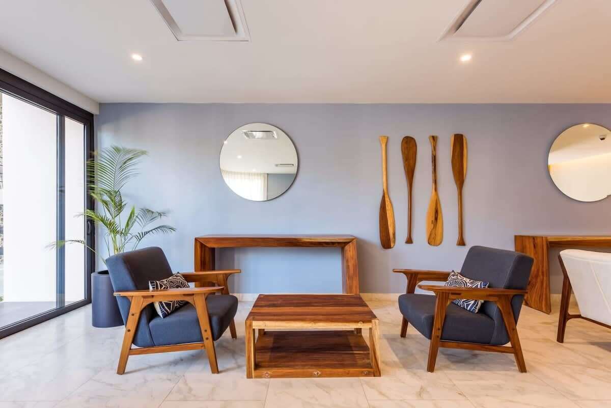 Una sala moderna con una mesa de centro de madera, dos sillones azules, un espejo redondo y palas de madera en la pared.