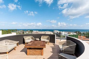 A rooftop terrace with wicker furniture, a wooden table, and a view of the ocean and cityscape. - Ground-Floor Studio w Rooftop Pool Near Beach (Playa del Carmen)