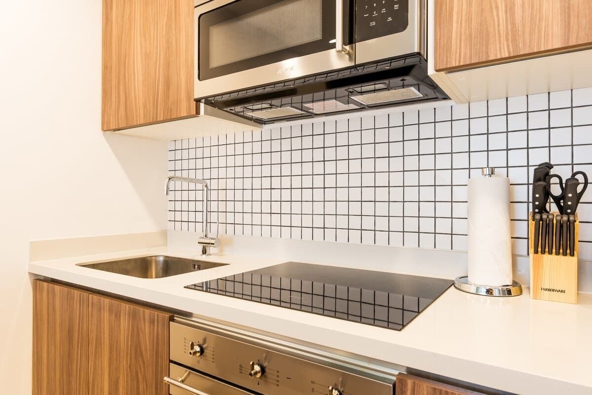 Une cuisine moderne équipée d’un four à micro-ondes en acier inoxydable, d’un four encastré et d’une plaque de cuisson à induction noire. On y trouve un évier blanc avec un robinet, ainsi qu’un bloc à couteaux en bois posé sur le plan de travail.