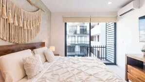 A bedroom with a large bed, a wooden headboard, a wall-mounted macrame piece, a bedside lamp, and a view of a building through a glass door. - 1BR Condo w Rooftop Pool Steps from Beach & 5th Ave (Playa del Carmen)