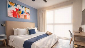 Camera d'albergo con un letto grande, una scrivania, una sedia, una finestra con tende e un quadro astratto dai colori vivaci appeso al muro.