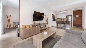 A modern living room with a sofa, a coffee table, and a TV. There is a kitchen area with a dining table and chairs. - Rooftop Pool & Gym Near 5th Avenue Activities (Playa del Carmen)