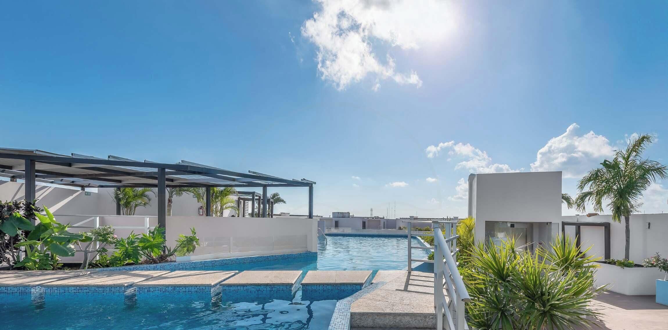 A rooftop swimming pool with a covered area, surrounded by plants and a clear sky.