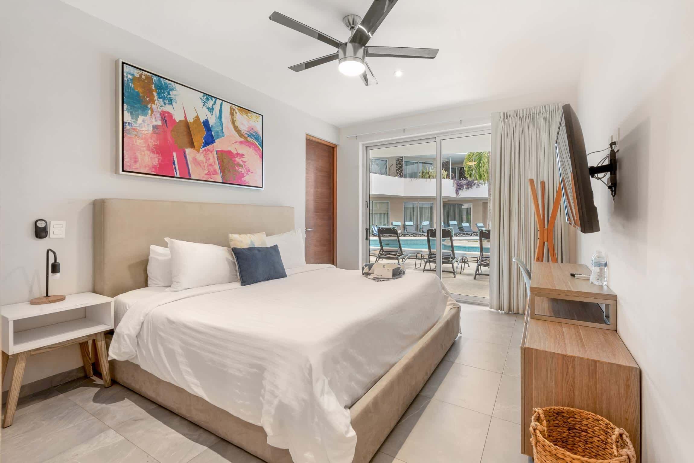 A hotel room with a large bed, a ceiling fan, a TV mounted on the wall, and a view of a pool outside.