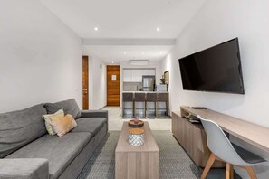 A modern living room with a grey sofa, a coffee table, a TV mounted on the wall, and a kitchen in the background. - Charming Beachside Condo w High-Speed WiFi (Playa del Carmen)