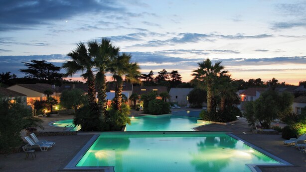 A large outdoor swimming pool with a curved design, surrounded by palm trees and lounge chairs. - CampingSpa - Confort 3 TV - 6p (La Couarde-sur-Mer)
