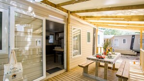 A wooden deck with a picnic table set up, a sliding glass door leading to a kitchen, and a small outdoor seating area. - CampingSpa - Confort 3 TV - 6p (La Couarde-sur-Mer)