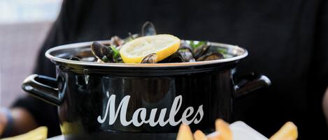 A black pot labeled "Moules" with mussels and lemon on a table, accompanied by a plate of fries.