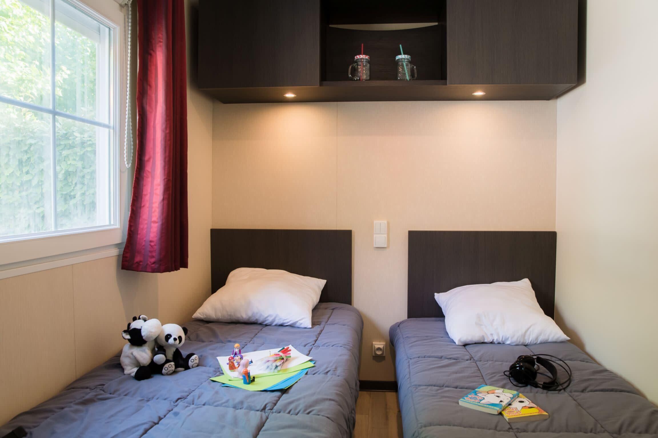 Two single beds with grey bedding, a window with red curtains, and a shelf above the beds holding two mugs.