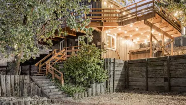 Une cabane rustique en bois, dotée d’un escalier en bois menant à un balcon, entourée d’arbres et d’une clôture en bois.