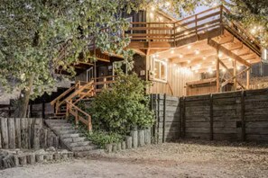 A rustic wooden cabin with a wooden staircase leading to a balcony, surrounded by trees and a wooden fence.