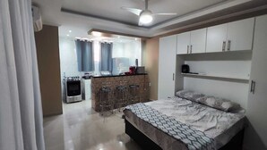 A modern bedroom with a large bed, a built-in wardrobe, and a kitchenette area. - Complete Studio 3 (Rio das Ostras)