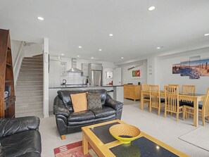 A modern living room with a black leather sofa, a wooden dining table with chairs, a kitchen in the background, and a staircase leading to another level.