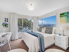 A bedroom with a large window, a bed with patterned pillows, a bedside table, and a desk with a chair. - Panoramic Views on Paku - Tairua Family Retreat (Tairua)