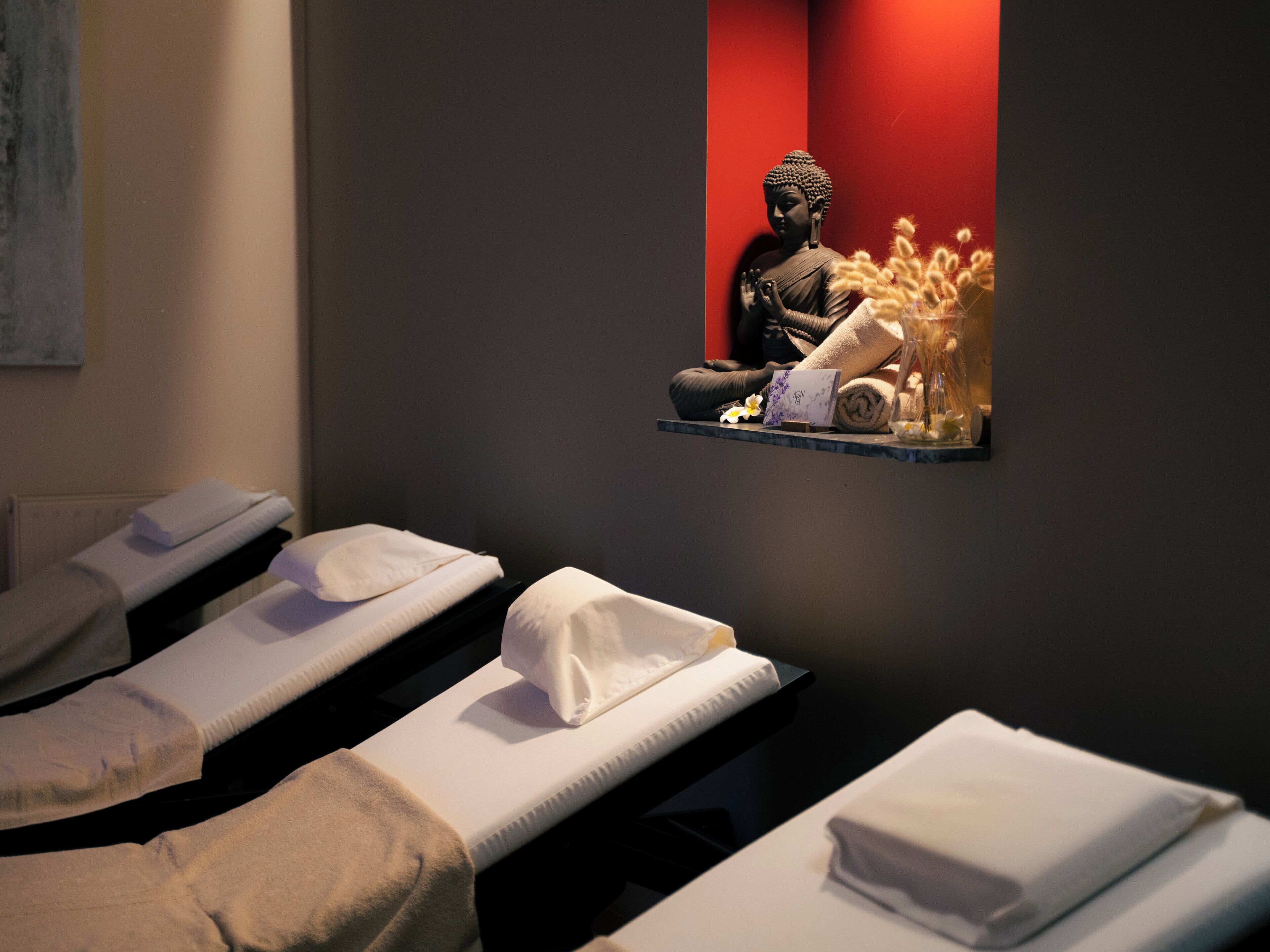 Una stanza spa con lettini per massaggi, una statua di Buddha e un piccolo ripiano con asciugamani e un vaso di fiori.