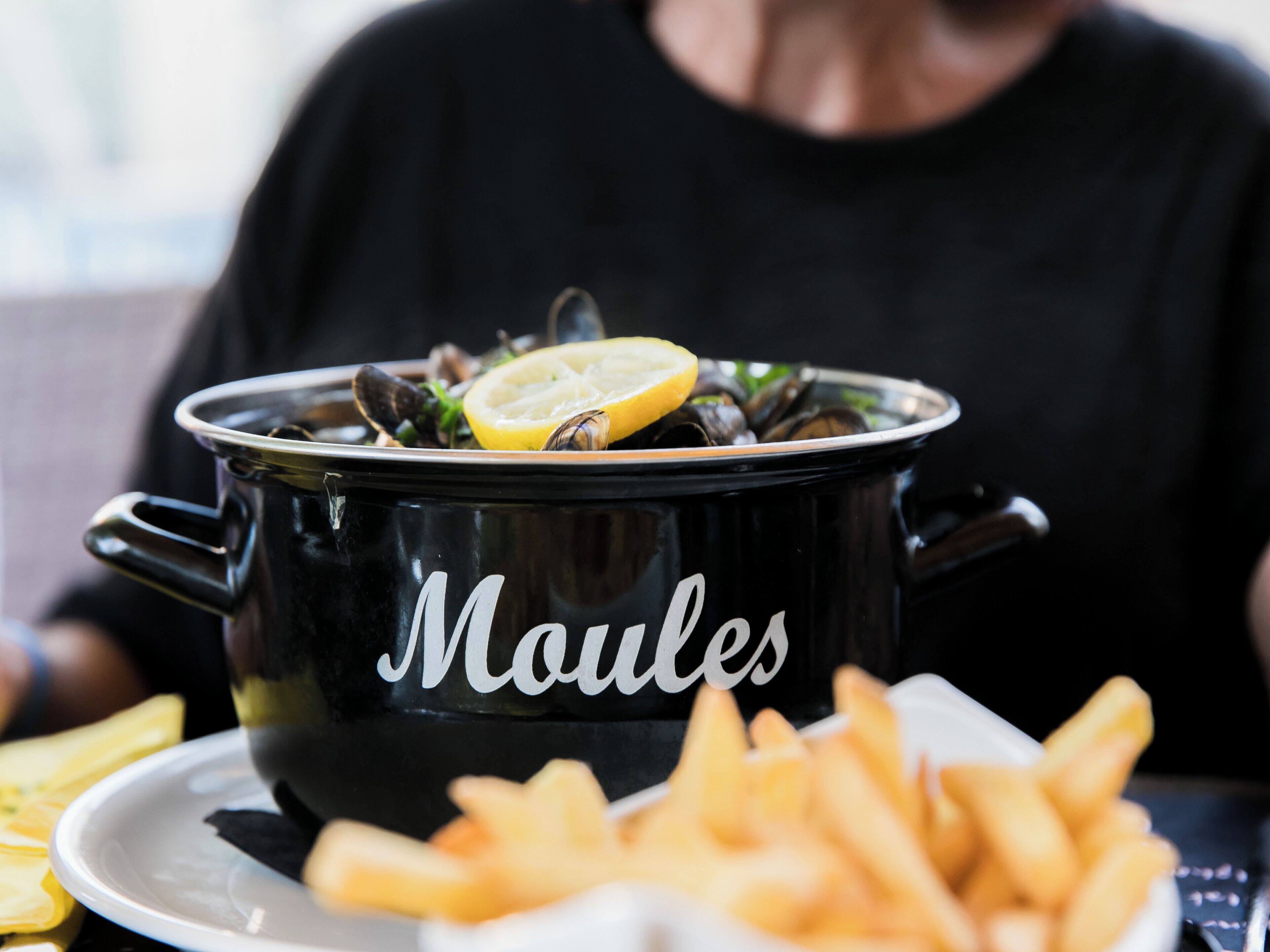 Une marmite noire étiquetée « Moules », contenant des moules et des tranches de citron, est posée sur une table, accompagnée d’une assiette de frites.