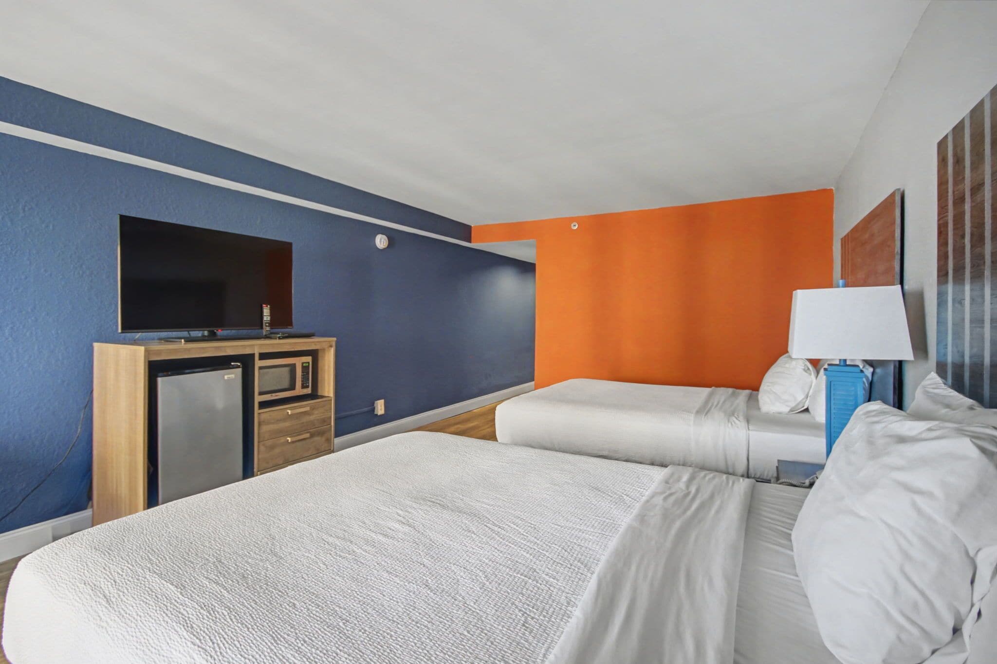 Une chambre d’hôtel avec deux lits, un bureau avec une télévision, un four à micro-ondes et une lampe bleue.