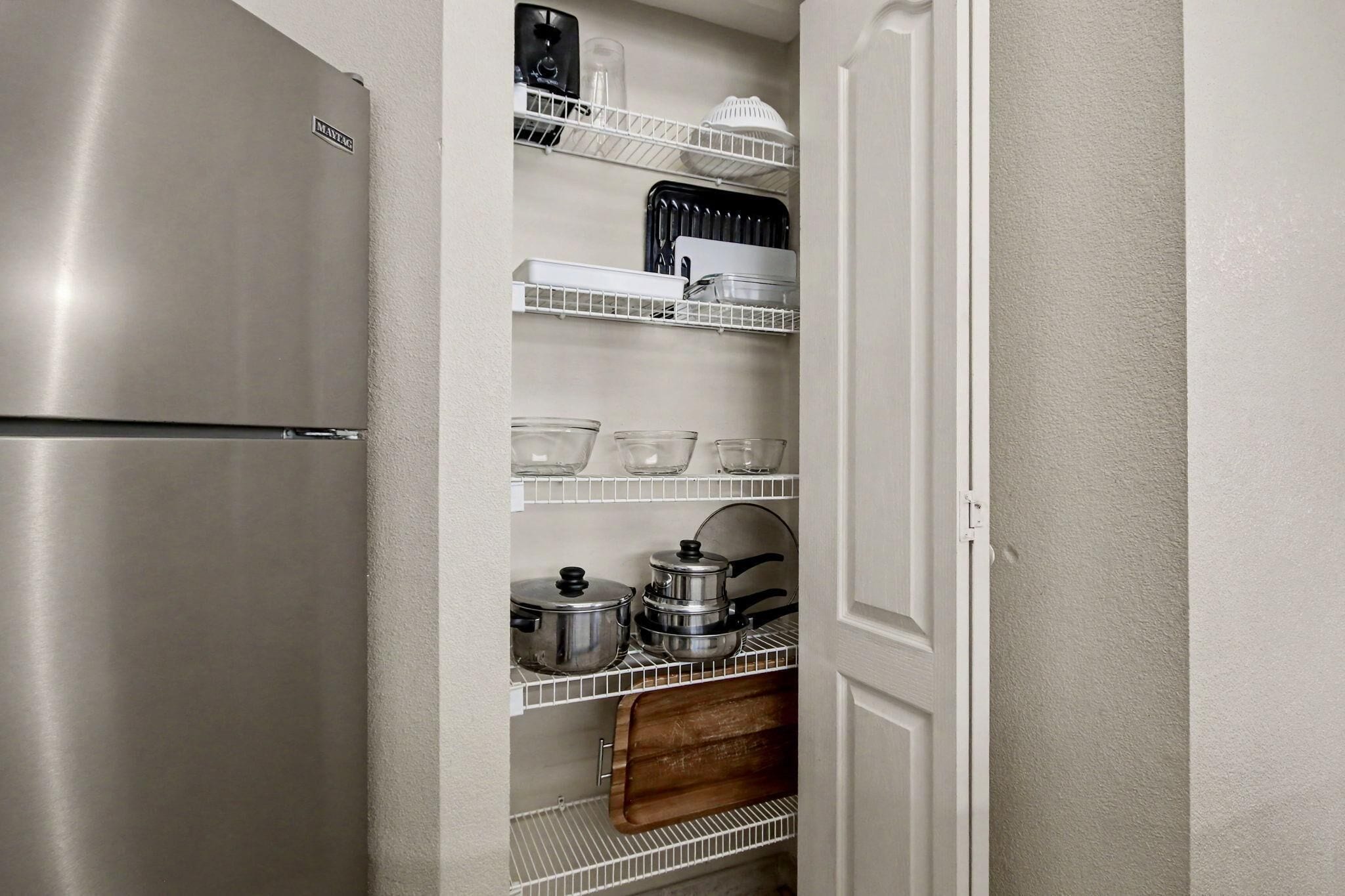 A kitchen with a stainless steel refrigerator, white shelving with dishes, glasses, pots, and a wooden cutting board.
