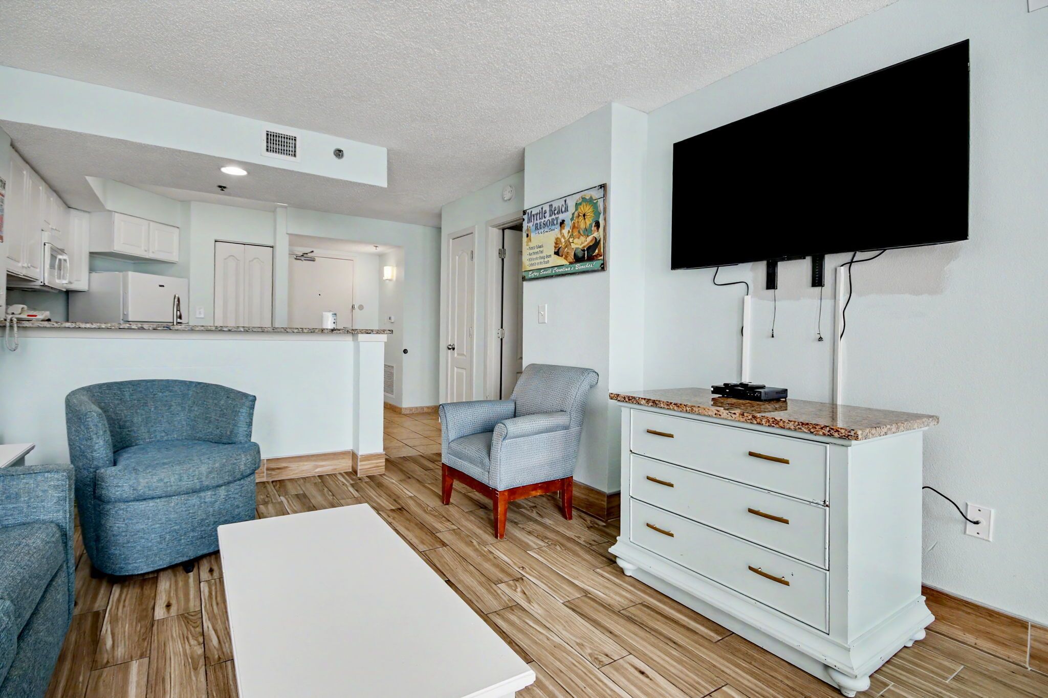Una cocina moderna con armarios blancos, un comedor con mesa blanca y una sala de estar con un sillón azul y un mueble blanco.