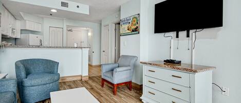 Una cocina moderna con armarios blancos, un comedor con mesa blanca y una sala de estar con un sillón azul y un mueble blanco.