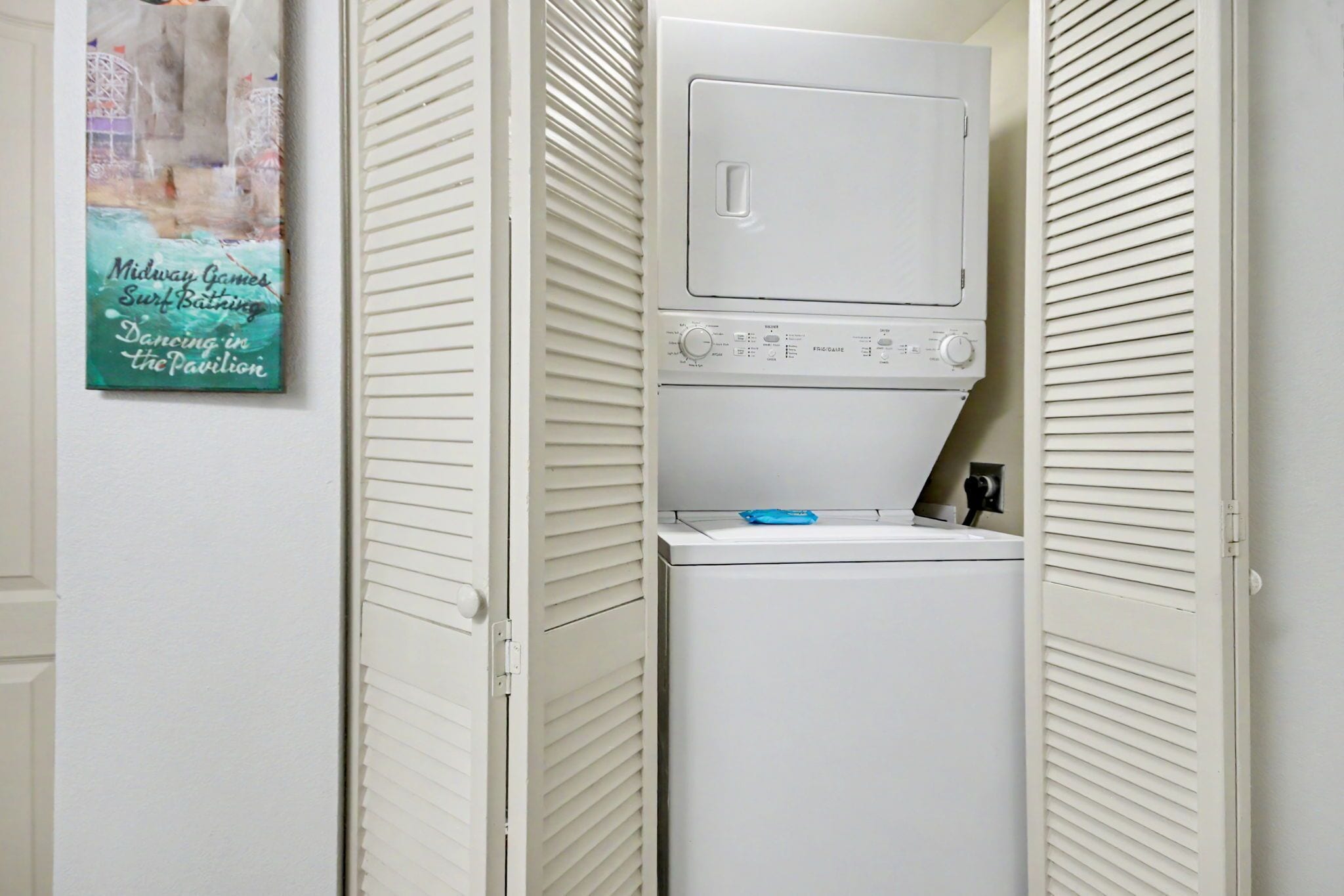 A laundry room with a stacked washer and dryer, a poster on the wall, and white louvered doors.