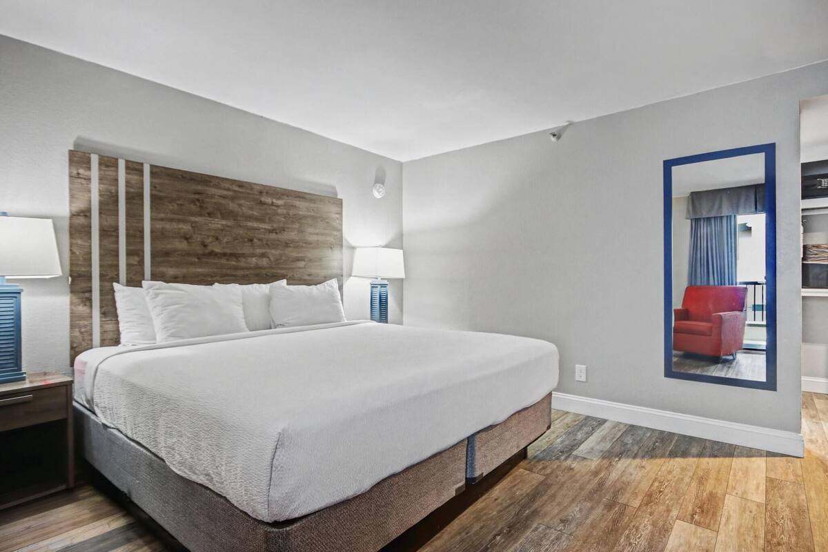 Una habitación de hotel con una cama grande, cabecera de madera, mesita de noche con lámpara, espejo en la pared y una silla roja en el reflejo.