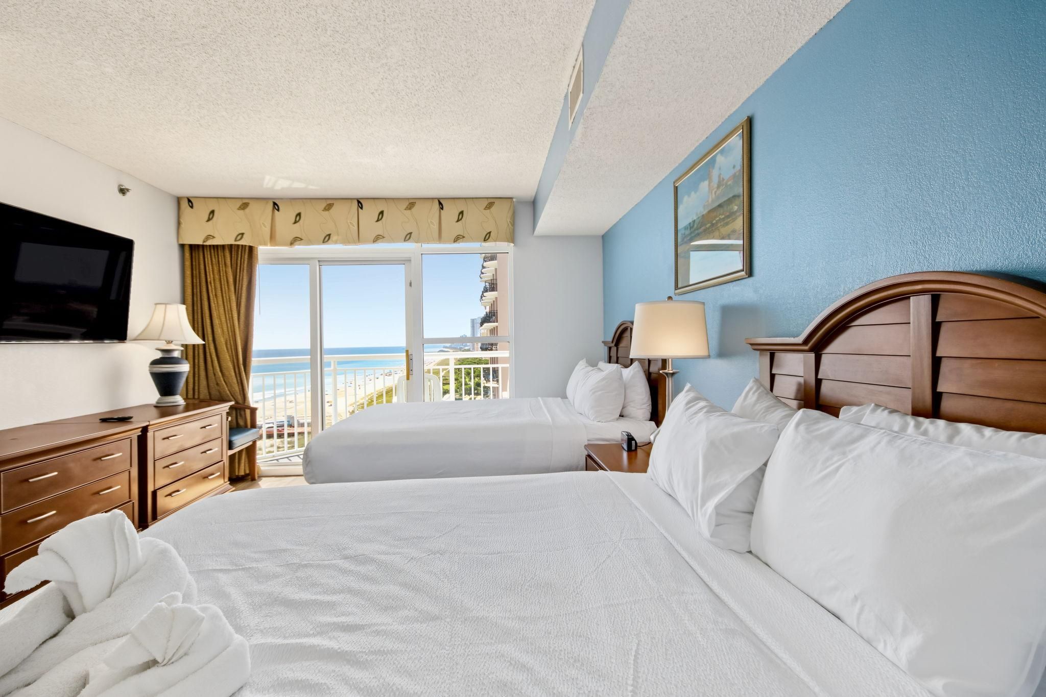 A hotel room with two beds, a TV, a desk, and a balcony with a view of the beach.