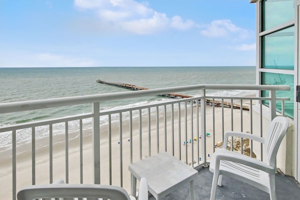 Un balcon avec vue sur la plage et l’océan, une table blanche et deux chaises blanches.