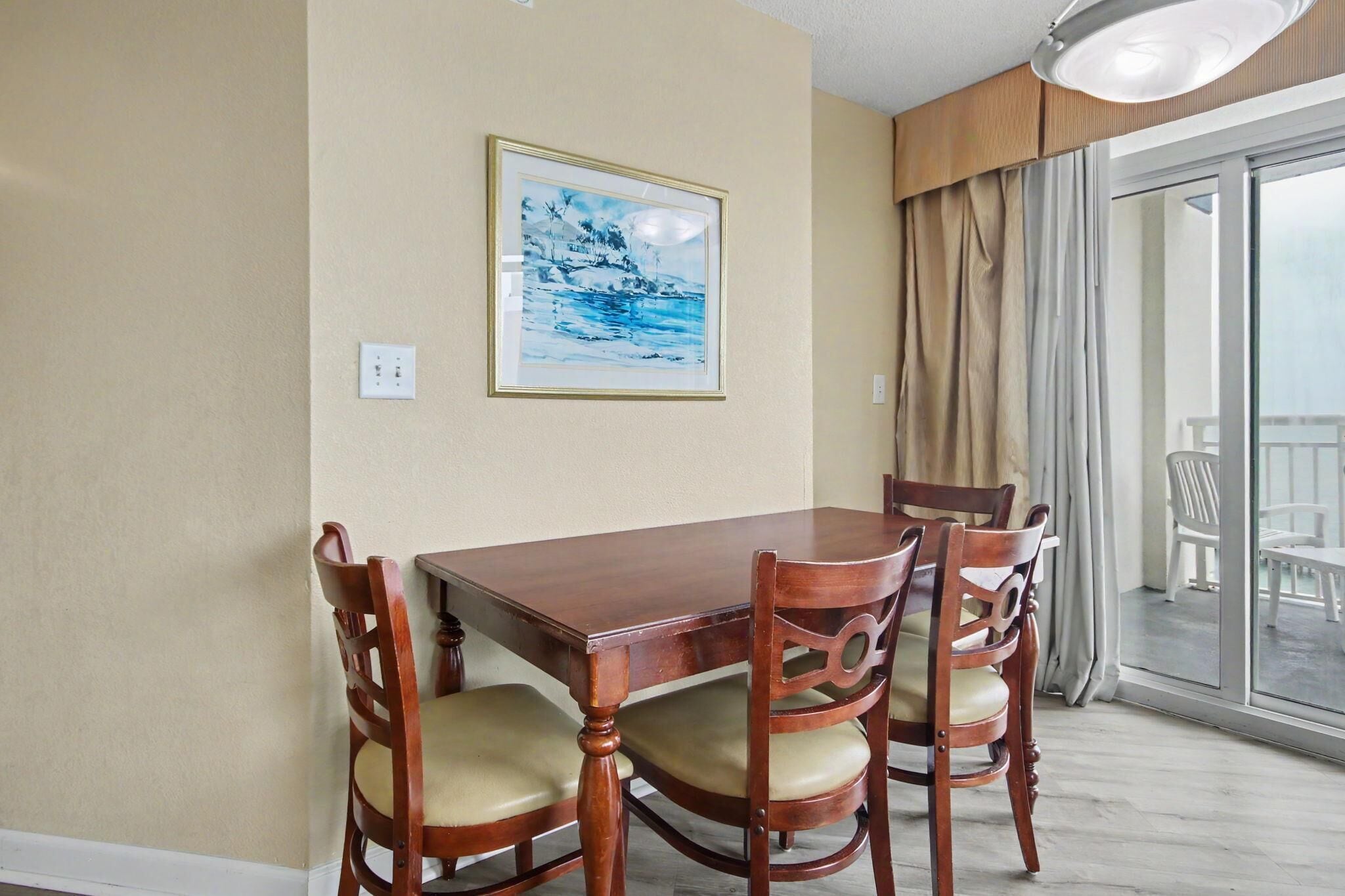 A dining area with a wooden table and chairs, a framed picture on the wall, and a view through a glass door.