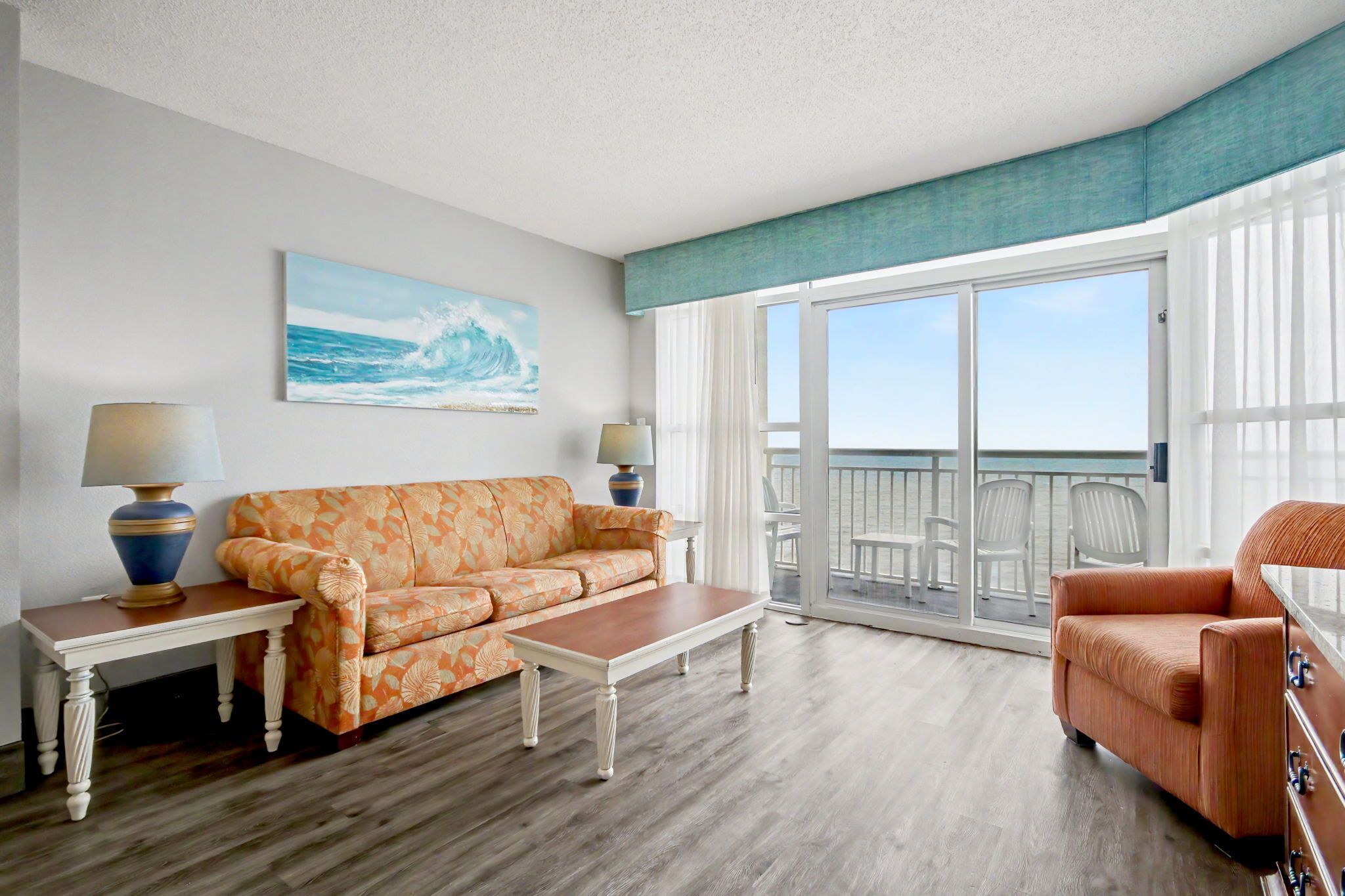 A living room with a sofa, coffee table, and armchair, a painting of the ocean, and a balcony with chairs.
