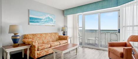 A living room with a sofa, coffee table, and armchair, a painting of the ocean, and a balcony with chairs.
