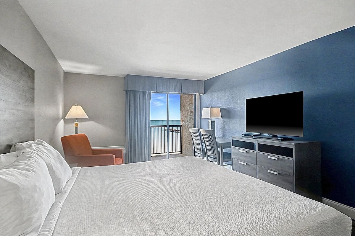 Habitación de hotel con una cama grande, televisión, escritorio con cajones, una silla, lámpara y vista al cielo a través de una ventana.