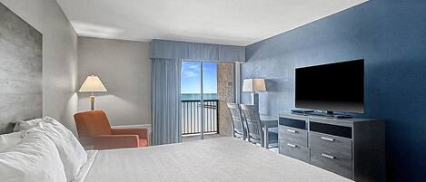 A hotel room with a large bed, a TV, a desk with drawers, a chair, a lamp, and a view of the sky through a window.
