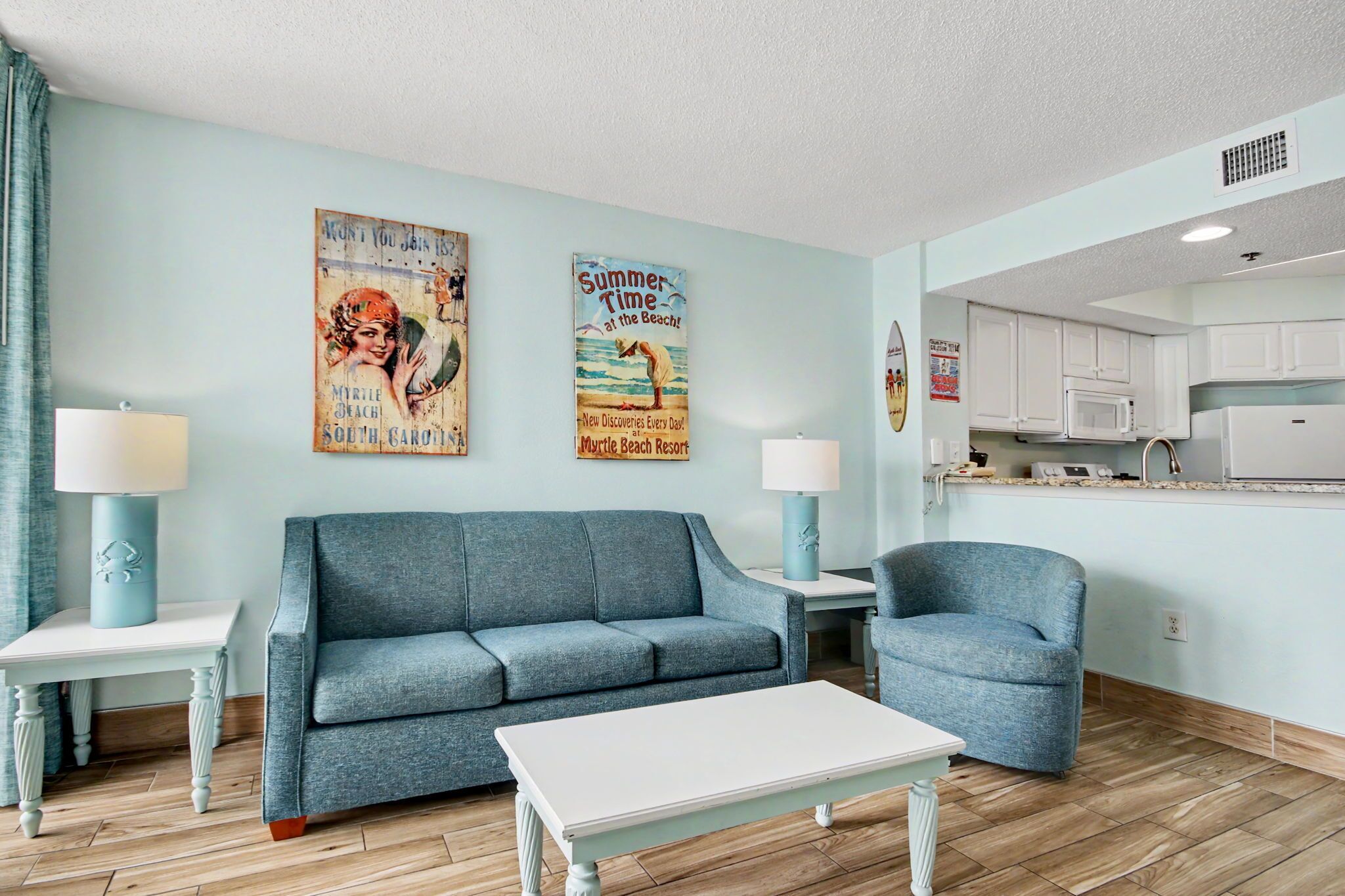 A living room with a sofa, armchair, and coffee table, a kitchen with white cabinets and appliances, and two wall posters.