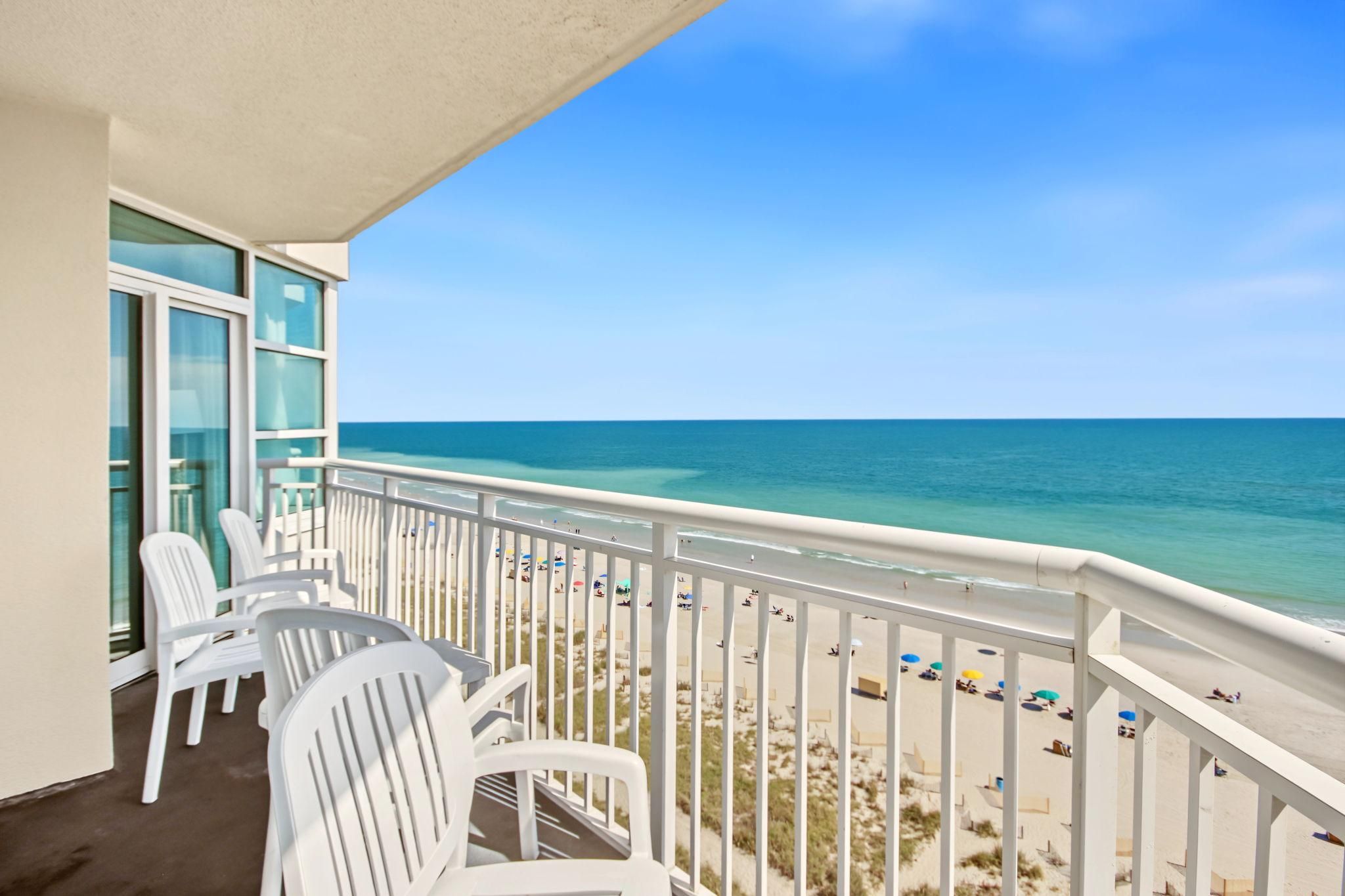 A balcony with white chairs, a view of the beach, and a clear sky.
