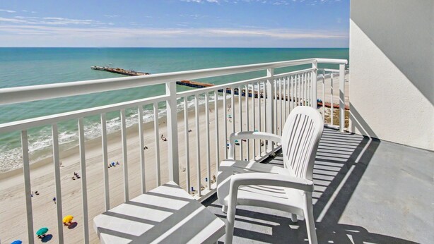 A balcony with white railings overlooking a beach with people and a pier. - Carolinian Beach Resort (Myrtle Beach)