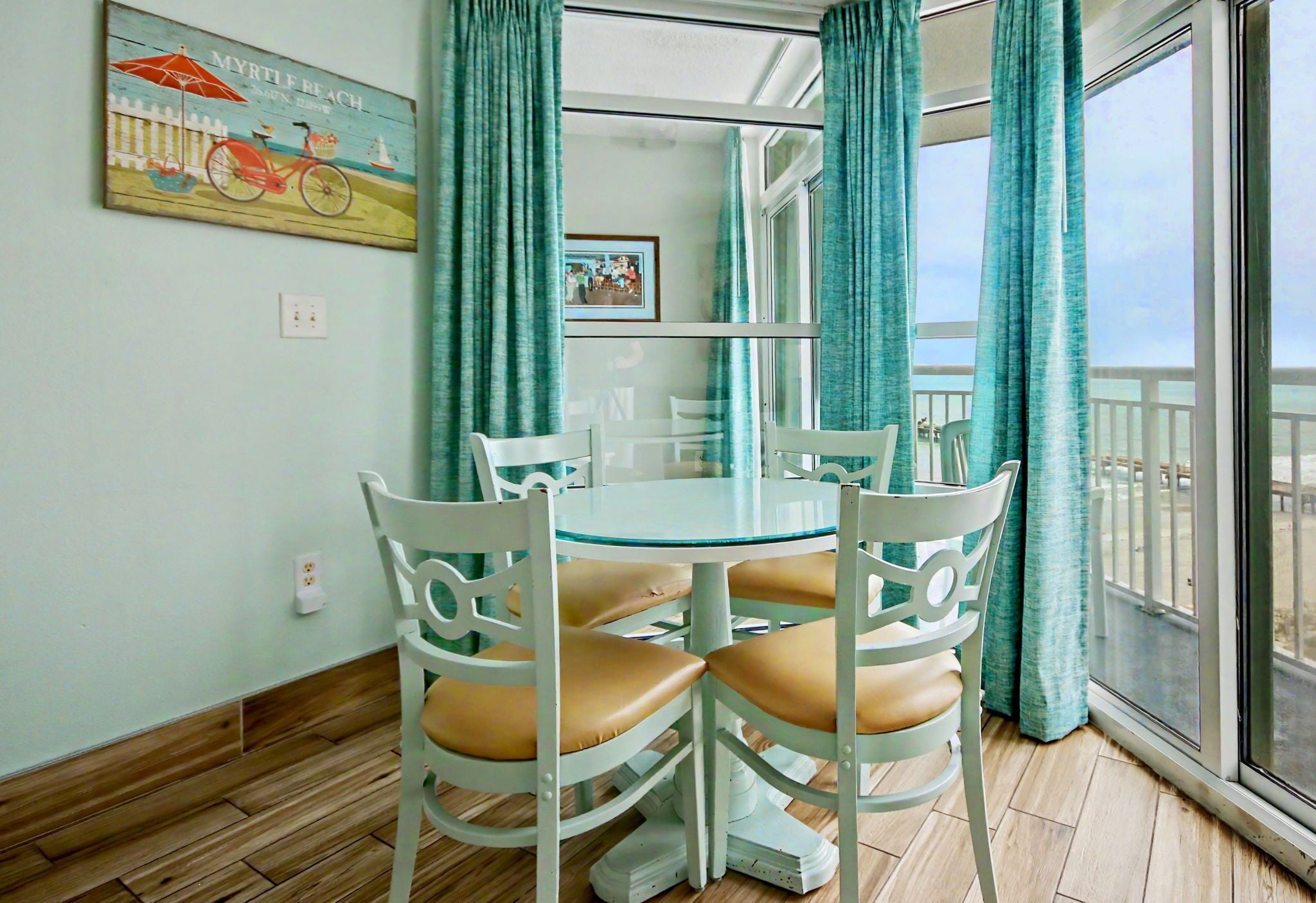 A room with a round glass table, white chairs, teal curtains, a wooden floor, and a beach-themed painting on the wall.